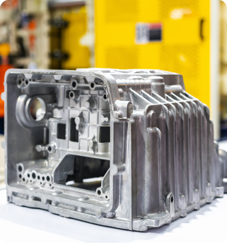 A detailed, metallic automotive engine part displayed on a workbench, with yellow industrial machinery blurred in the background.
